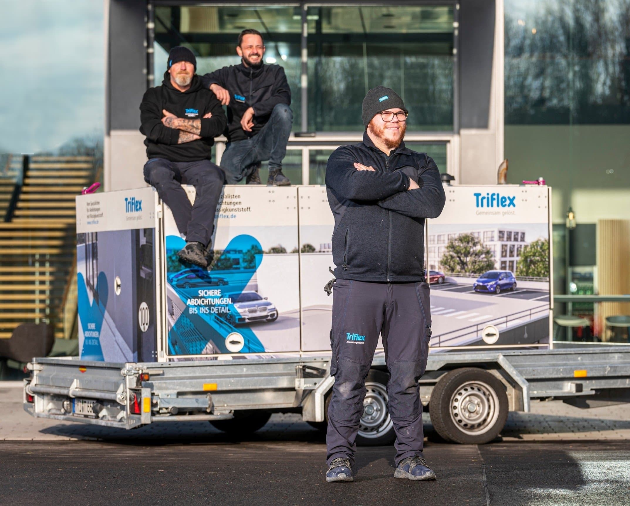 Three skilled workers in work clothes are standing in front of a modern commercial building with a glass facade on an outdoor construction site.
