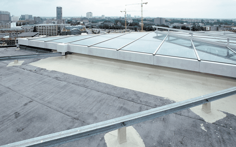 Flachdach mit frischer Dachabdichtung neben großflächiger Glasdachkonstruktion, aufgenommen über einer urbanen Skyline.