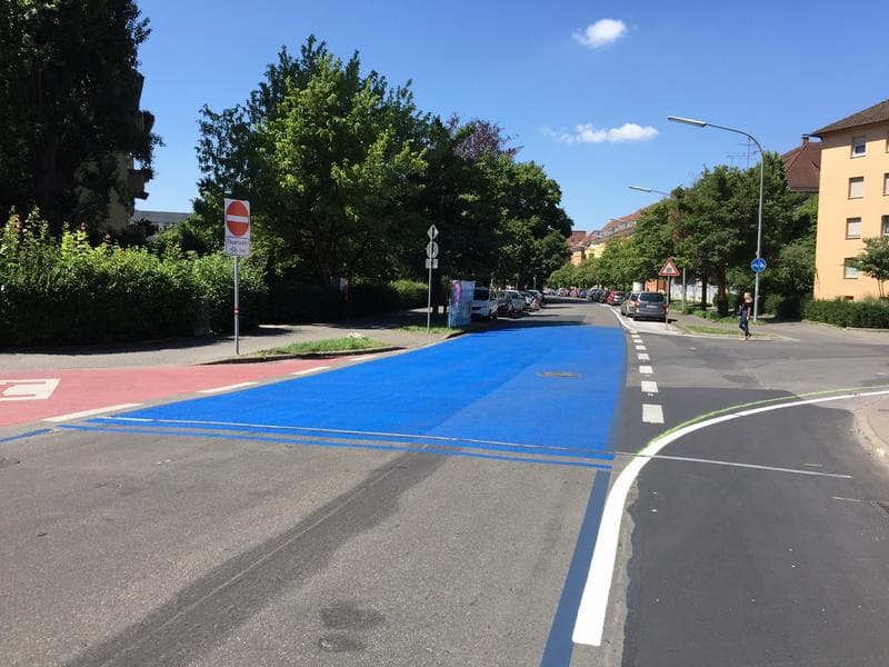 Straßenkreuzung mit deutlich markierter blauer Fahrradspur und rotem Radweg vor Wohnhäusern; gut sichtbare Fahrbahnmarkierungen zur Optimierung der Verkehrssicherheit.