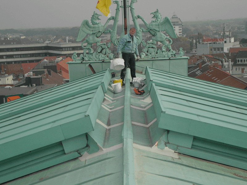 Handwerker bei Wartungsarbeiten auf einem großflächigen Kupferdach mit grüner Patina, Stadtansicht im Hintergrund.