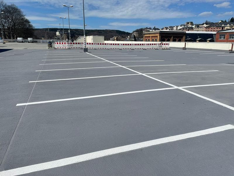 Frisch markierter Parkplatz mit grauem Industrieboden und weißen Parkplatzlinien vor Gewerbegebäuden unter blauem Himmel.