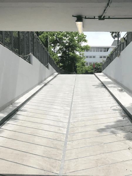 Betonrampe in einer modernen Tiefgaragenzufahrt mit weißen Seitenwänden und Geländern, im Hintergrund grüne Bäume und ein Bürogebäude bei Tageslicht.